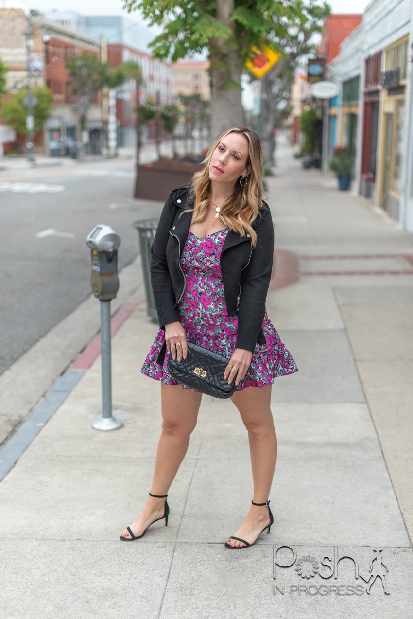 Shein Floral Dresses by popular LA fashion blog, Posh in Progress: image of of a woman standing outside on a sidewalk in front of some shops and wearing a purple floral print ruffle hem Shein dress, leather jacket, black ankle strap sandals, and holding a black clutch. 